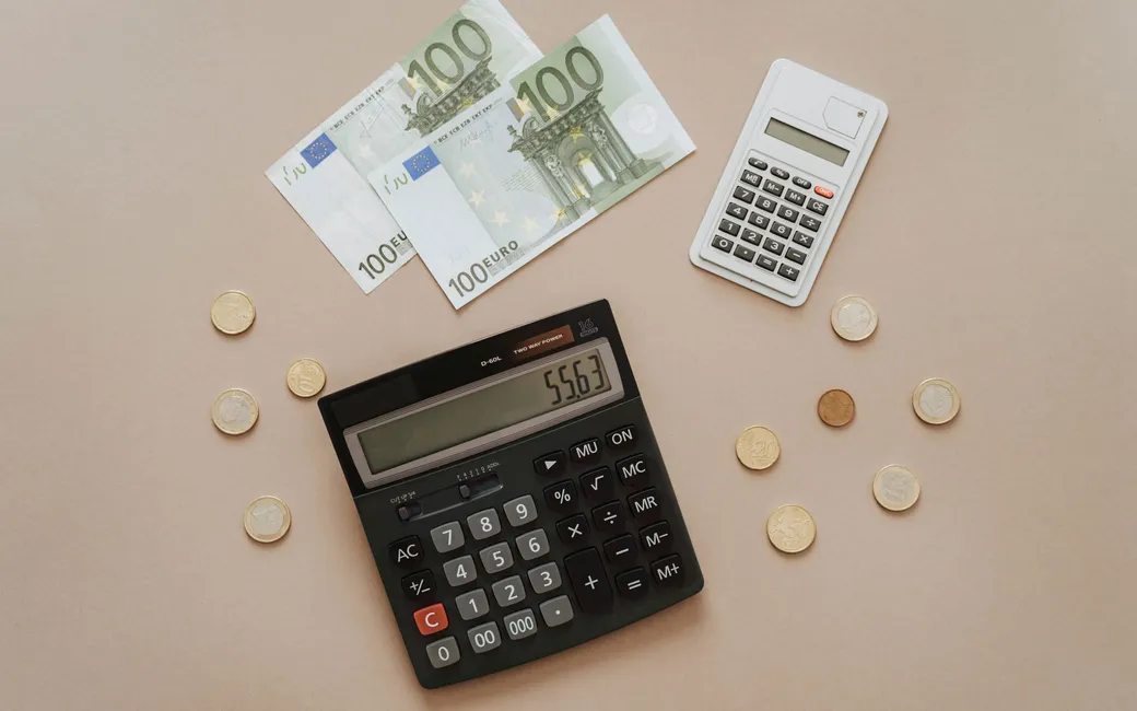 Twee rekenmachines, twee biljetten van honderd euro en losse munten op een beige ondergrond