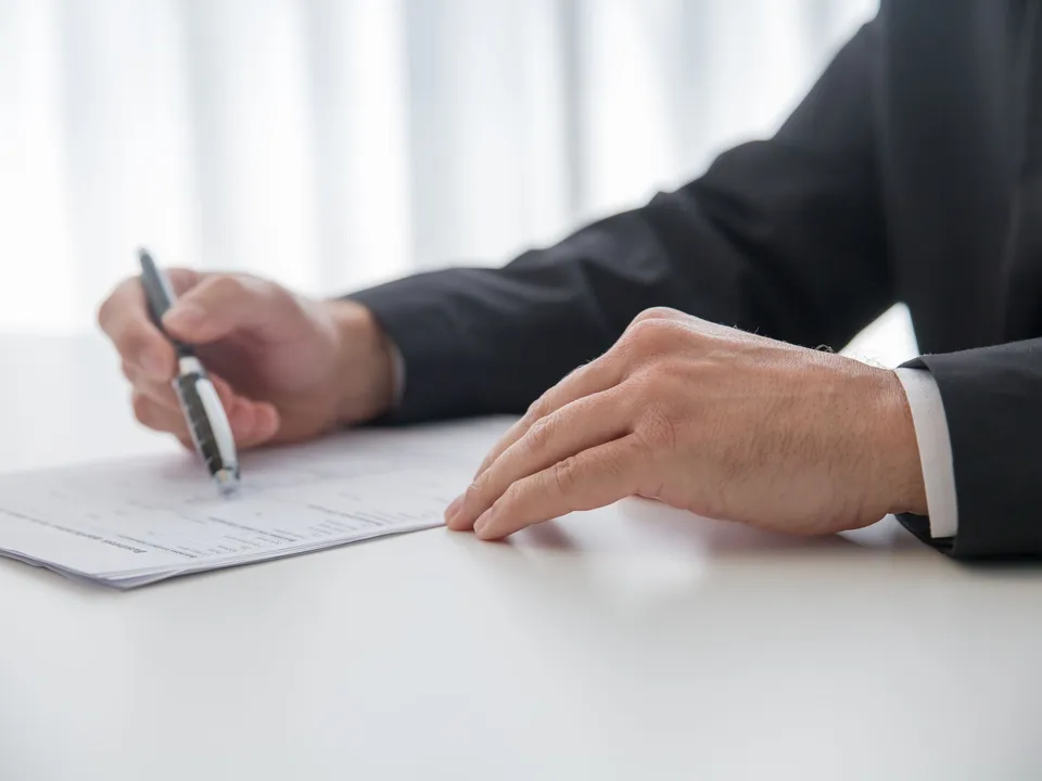 Handen van iemand in donker pak vullen een formulier in aan een wit bureau