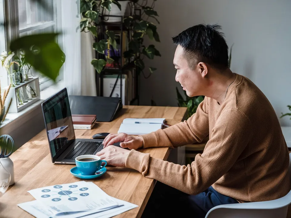 Man werkt aan een laptop aan een houten bureau bij het raam, tussen planten