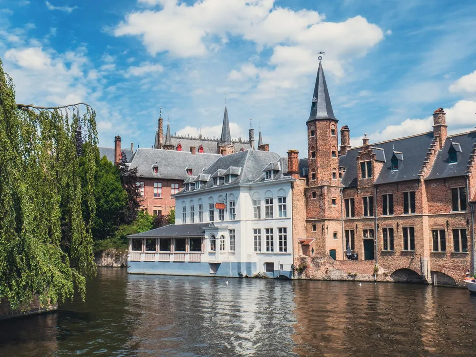 Historische bakstenen gevels en een treurwilg langs een Brugse gracht onder blauwe wolkenlucht